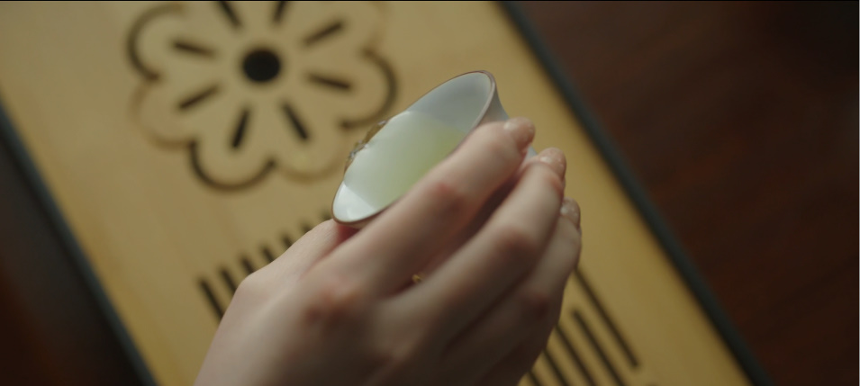 A hand holding a small teacup over a tea tray, showing a formal tea ceremony detail in a Korean drama scene.